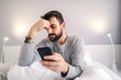 © Dusan Petkovic - Depressed lonely caucasian bearded man sitting on bed in bedroom at night, holding his head and using smart phone for online chatting. Problems in relationship.