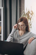 © Kuz Production - Serious dark-haired woman in eyeglasses using laptop for remote work at home. Attractive girl in casual outfit looking at computer screen while sitting on sofa