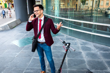 Wall Mural - Smiling businessman with electric scooter standing in front of modern business building talking on phone.