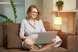 © Roman Tyukin - A woman in a dress sitting on the couch with legs crossed works remotely on a laptop in her apartment. A girl with braces watching a webinar. A female student in glasses listening to an online lecture