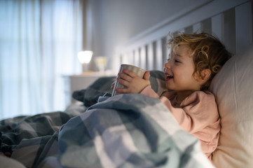 Wall Mural - Small sick toddler girl lying in bed indoors at home, holding cup.