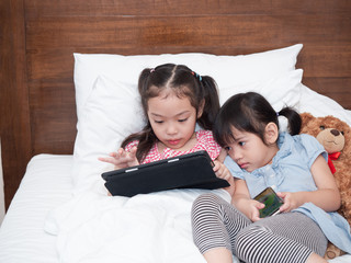 Two Asian sibling girl looking at tablet and smartphone for playing game , looking cartoon or learning online on the bed at home.