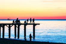 The Fishermen On The Pier Fishing Against The Background Of The Sea Sunset