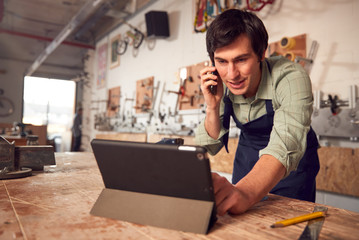 Male Business Owner In Workshop Using Digital Tablet And Making Call On Mobile Phone