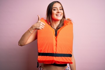 Young beautiful brunette woman wearing orange lifejacket over isolated pink background doing happy thumbs up gesture with hand. Approving expression looking at the camera showing success.