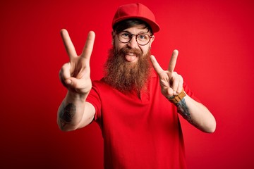 Wall Mural - Young handsome delivery man wearing glasses and red cap over isolated background smiling with tongue out showing fingers of both hands doing victory sign. Number two.