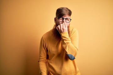 Canvas Print - Handsome Irish redhead man with beard wearing glasses over yellow isolated background smelling something stinky and disgusting, intolerable smell, holding breath with fingers on nose. Bad smell