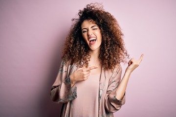 Young beautiful woman with curly hair and piercing wearing casual pink t-shirt smiling and looking at the camera pointing with two hands and fingers to the side.