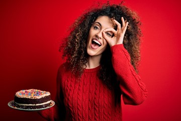 Young beautiful woman with curly hair and piercing holding plate with birthday cake with happy face smiling doing ok sign with hand on eye looking through fingers