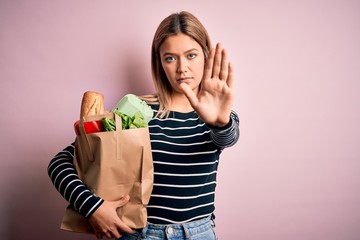 Sticker - Young beautiful woman holding paper bag with purchase over isolated pink background with open hand doing stop sign with serious and confident expression, defense gesture