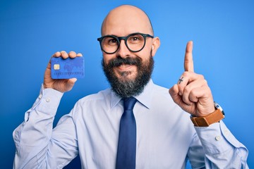 Poster - Handsome bald business man with beard holding credit card over isolated blue background surprised with an idea or question pointing finger with happy face, number one