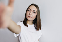 Good-looking European Woman Making Selfie On White Background. Inspired Young Lady With Dark Hair Taking Picture Of Herself With Serious Face Expression.