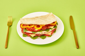 Wall Mural - fresh sandwich with salami, pita, vegetables and cheese served on plate near golden fork and knife on green background