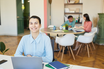 Wall Mural - Cheerful young adult businesswoman sitting at office desk with laptop on it smiling at camera, her colleagues working behind her