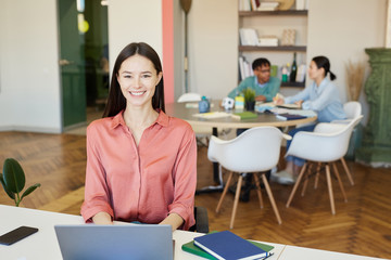 Wall Mural - cheerful young woman wearing pink blouse sitting at office desk looking at camera smiling, her colle