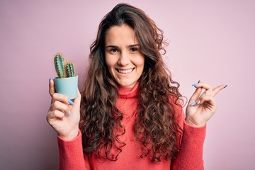 Sticker - Young beautiful woman with curly hair holding small cactus over isolated pink background very happy pointing with hand and finger to the side