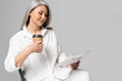 © LIGHTFIELD STUDIOS - smiling asian woman with grey hair reading business newspaper with coffee to go isolated on grey