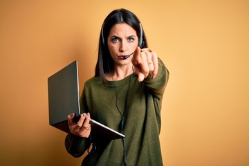 Wall Mural - Young operator woman with blue eyes working on call center using computer laptop and headset pointing with finger to the camera and to you, hand sign, positive and confident gesture from the front