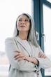© LIGHTFIELD STUDIOS - corporate asian businesswoman with crossed arms and grey hair standing in grey suit and eyeglasses in office