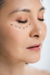 © LIGHTFIELD STUDIOS - asian woman with closed eyes and plastic surgery lines on face isolated on grey