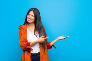 young pretty latin woman smiling, feeling happy, carefree and satisfied, pointing to concept or idea on copy space on the side against flat wall