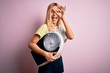 Young beautiful blonde sporty woman on diet holding weight machine over pink background with happy face smiling doing ok sign with hand on eye looking through fingers