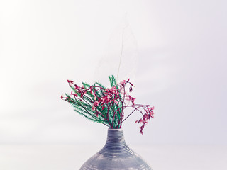 Wall Mural - Flower composition. A bouquet of dried plants (flowers or leaves) is in a small striped vase on a white wooden table. Behind is a white wall with shadows of plants. Copy space.