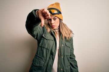 Poster - Young brunette skier woman wearing snow clothes and ski goggles over white background looking unhappy and angry showing rejection and negative with thumbs down gesture. Bad expression.