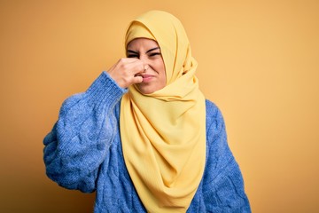 Poster - Young beautiful brunette muslim woman wearing arab hijab over isolated yellow background smelling something stinky and disgusting, intolerable smell, holding breath with fingers on nose. Bad smell