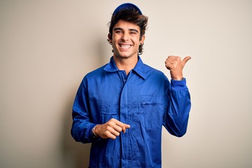 Wall Mural - Young mechanic man wearing blue cap and uniform standing over isolated white background smiling with happy face looking and pointing to the side with thumb up.