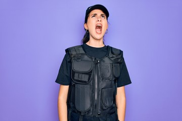 Canvas Print - Young police woman wearing security bulletproof vest uniform over purple background angry and mad screaming frustrated and furious, shouting with anger. Rage and aggressive concept.