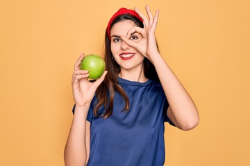 Poster - Young beautiful girl eating fresh organic healthy green apple over yellow background with happy face smiling doing ok sign with hand on eye looking through fingers