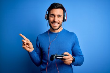 Young handsome gamer man with beard playing video game using joystick and headphones very happy pointing with hand and finger to the side