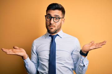 Sticker - Young handsome businessman wearing tie and glasses standing over yellow background clueless and confused with open arms, no idea concept.