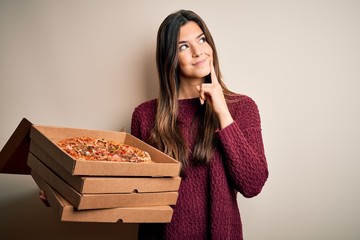 Poster - Young beautiful girl holding delivery boxes with Italian pizza standing over white background serious face thinking about question, very confused idea