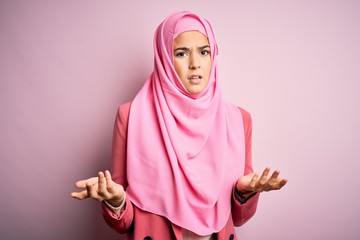 Canvas Print - Young beautiful girl wearing muslim hijab standing over isolated pink background clueless and confused with open arms, no idea concept.