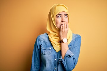 Poster - Young beautiful girl wearing muslim hijab standing over isolated yellow background looking stressed and nervous with hands on mouth biting nails. Anxiety problem.