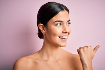 Canvas Print - Young beautiful girl standing over isolated pink background pointing and showing with thumb up to the side with happy face smiling