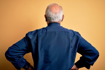 Grey haired senior man wearing casual blue shirt standing over yellow background standing backwards looking away with arms on body