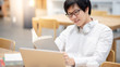 © zephyr_p - Young Asian man university student with glasses reading book while using laptop computer for online study course in college library. E-learning and doing research on internet. Education concept