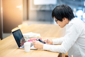 Wall Mural - Asian man university student with glasses and headphones using laptop computer and smartphone for online study course in college library. E-learning and doing research on internet. Education concept