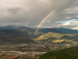 Fototapeta Tęcza - A beautiful, aerial view of a rainbow streaming through dark clouds over the green hills surrounding the village of Sarangkot near Pokhara in Nepal.