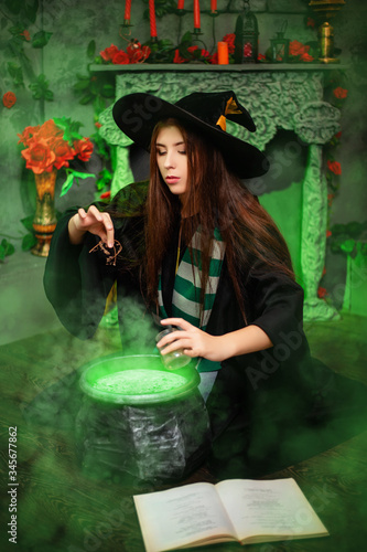 Fotomural Attractive girl in the image of a witch dressed in a black hat and mantle throws an ingredient into a cauldron with an upcoming potion, sitting in a magical, green smoke