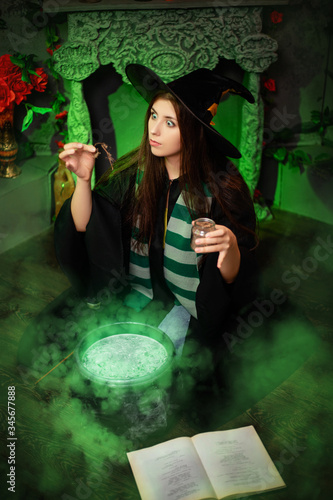 Fotografía Attractive girl in the image of a witch dressed in a black hat and mantle throws an ingredient into a cauldron with an upcoming potion, sitting in a magical, green smoke