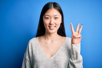 Wall Mural - Young beautiful asian woman wearing casual sweater standing over blue isolated background showing and pointing up with fingers number three while smiling confident and happy.