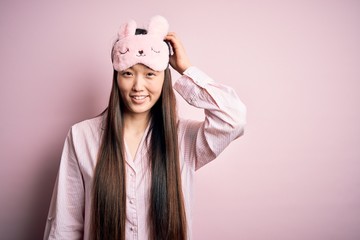 Sticker - Young asian woman wearing pajama and sleep mask over pink isolated background confuse and wonder about question. Uncertain with doubt, thinking with hand on head. Pensive concept.