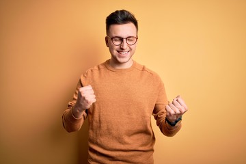 Wall Mural - Young handsome caucasian man wearing glasses and casual winter sweater over yellow background excited for success with arms raised and eyes closed celebrating victory smiling. Winner concept.