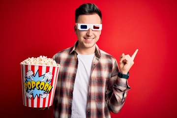 Wall Mural - Young handsome caucasian man wearing 3d movie glasses and eating popcorn cheerful with a smile on face pointing with hand and finger up to the side with happy and natural expression