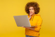Confused puzzled thoughtful woman with fluffy curly hair in urban style hoody holding laptop thinking over difficult question, pondering solution. indoor studio shot isolated on yellow background