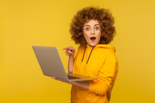 Amazed Emotional Bright Woman With Curly Hair In Urban Style Hoody Pointing Laptop Screen And Looking At Camera With Shocked Expression, Open Mouth. Indoor Studio Shot Isolated On Yellow Background